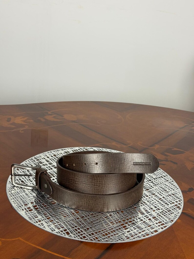 May include: A brown leather belt with a silver buckle and a textured surface. The belt is coiled on a white, patterned surface. The belt is a neutral color and could be worn with many outfits.