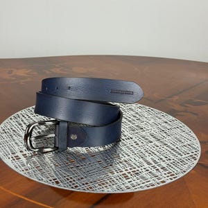 May include: A rolled-up navy blue leather belt with a silver-colored buckle. The belt is displayed on a decorative silver-colored plate. The belt has a dark blue suede detail near the buckle.