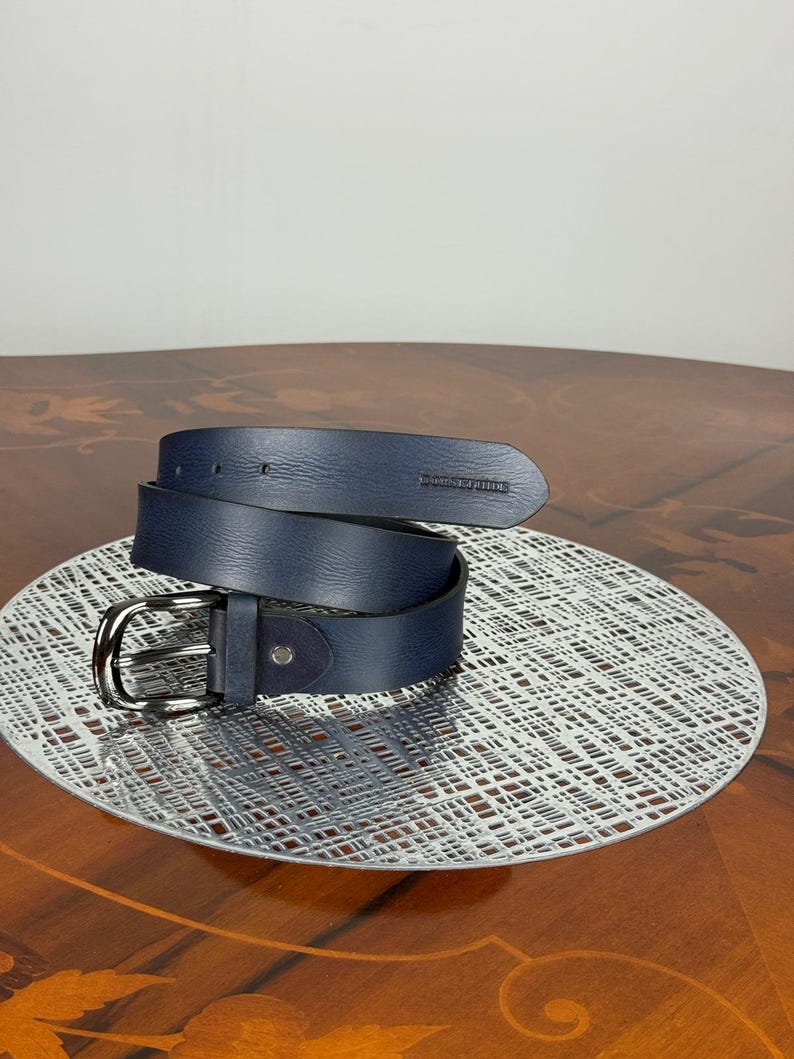 May include: A rolled-up navy blue leather belt with a silver-colored buckle. The belt is displayed on a decorative silver-colored plate. The belt has a dark blue suede detail near the buckle.