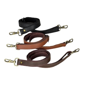 Puede incluir: Tres correas de cuero para bolsos en negro, marrón y beige. Cada correa tiene un cierre metálico en ambos extremos para facilitar la fijación a una bolsa. Las correas están enrolladas y colocadas sobre un fondo blanco.