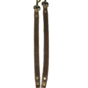 Op de afbeelding: Twee bruine leren riemen met messingkleurige hardware en een goudkleurige ring. De riemen hebben witte stiksels langs de randen en zijn aan de bovenkant verbonden met een draaihaak.