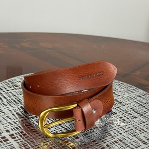 May include: A brown leather belt with a gold-colored buckle. The belt is rolled up and has the text "DOREEN MADE" embossed on the leather. The belt is on a silver patterned surface.