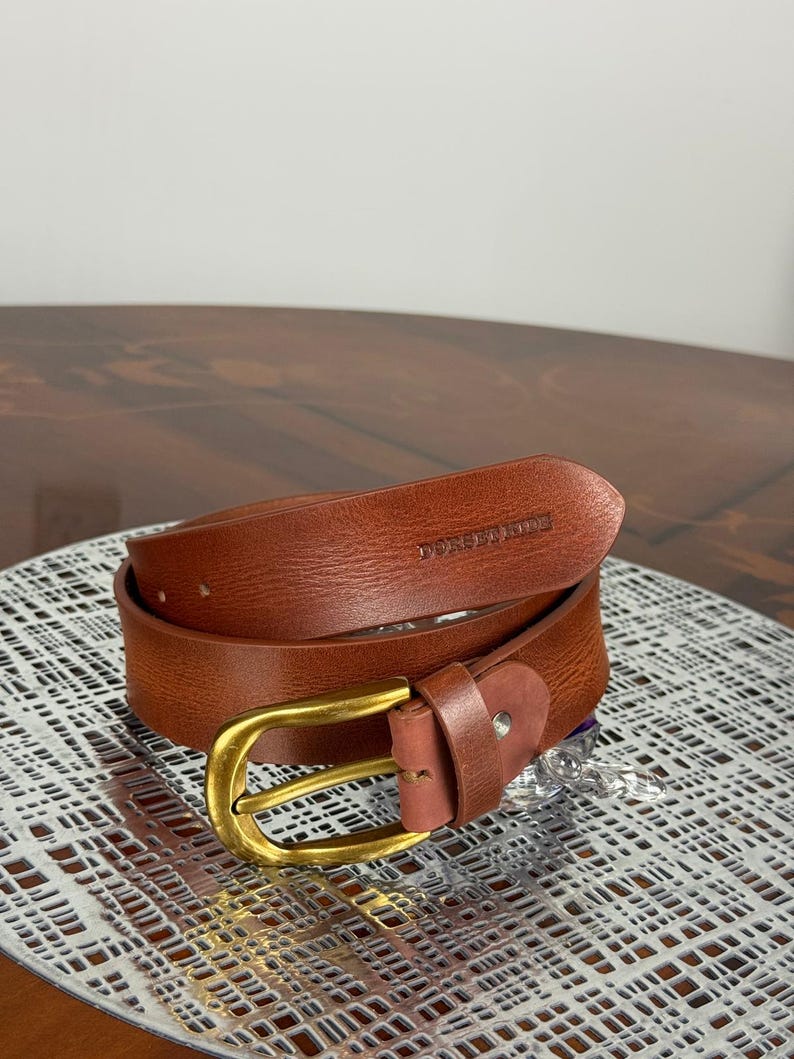May include: A brown leather belt with a gold-colored buckle. The belt is rolled up and has the text "DOREEN MADE" embossed on the leather. The belt is on a silver patterned surface.