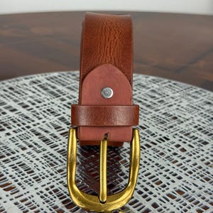 May include: A brown leather belt with a gold-colored buckle. The belt is made of a textured leather material and features a classic buckle design. The buckle is a shiny gold color and has a rectangular shape.