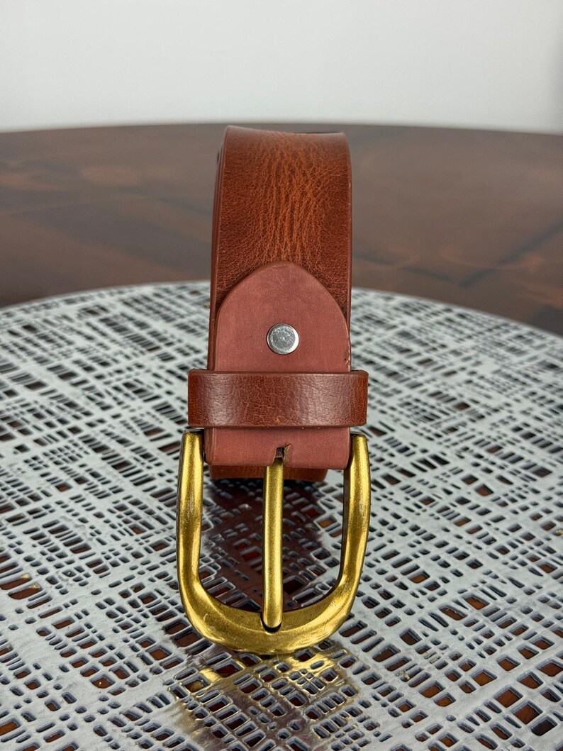 May include: A brown leather belt with a gold-colored buckle. The belt is made of a textured leather material and features a classic buckle design. The buckle is a shiny gold color and has a rectangular shape.