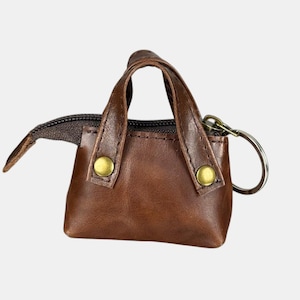 May include: A miniature brown leather handbag-style keyring with a zip closure. The bag features two small handles and is fastened with two gold-coloured buttons. A silver-coloured key ring is attached.