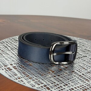 May include: A navy blue belt with a silver-colored buckle. The belt is made of a textured material and is rolled up on a decorative surface. The buckle is rectangular with a rounded edge.