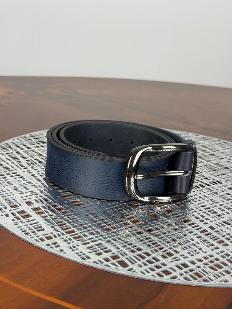 May include: A navy blue belt with a silver-colored buckle. The belt is made of a textured material and is rolled up on a decorative surface. The buckle is rectangular with a rounded edge.