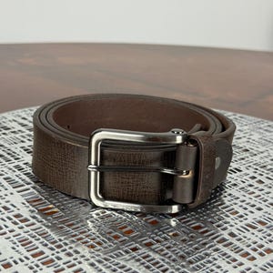 May include: A rolled-up brown leather belt with a silver-colored buckle. The belt has a textured surface and a classic design. The buckle is rectangular and has a single prong.