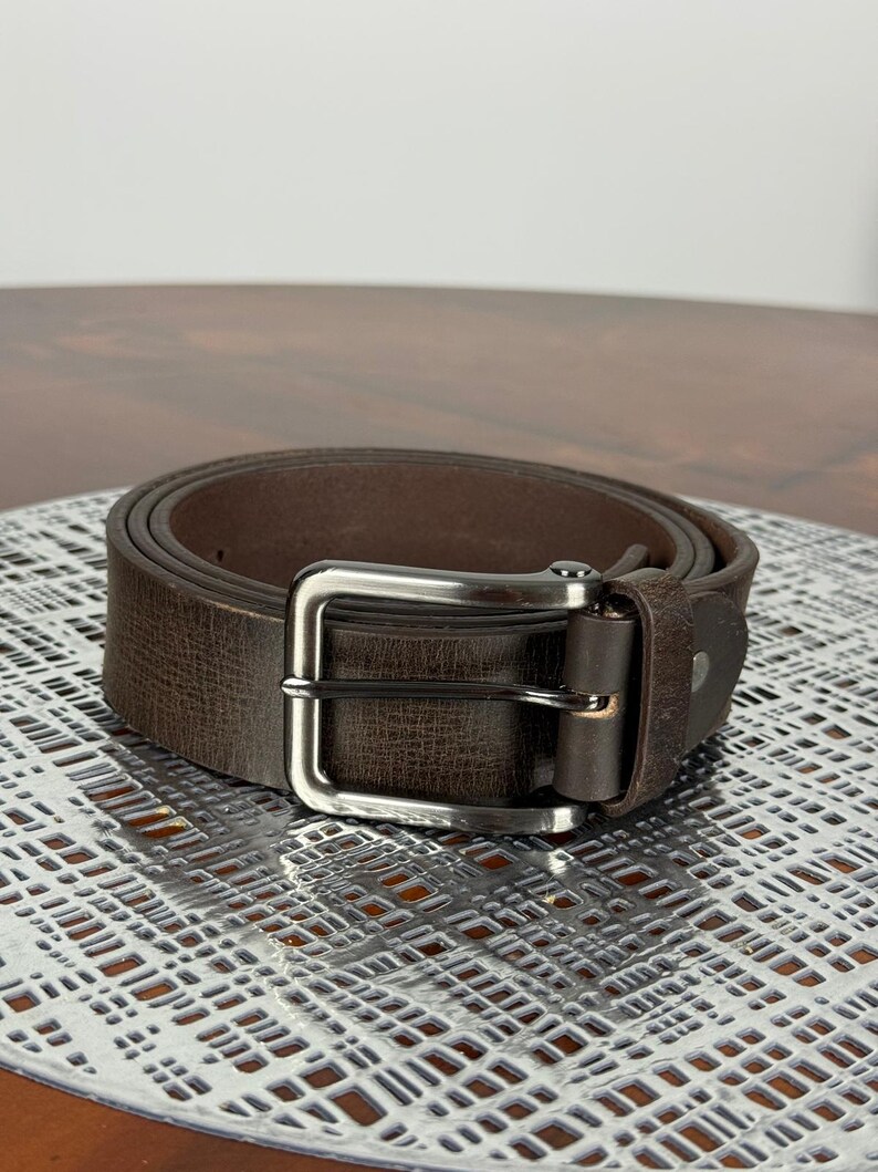 May include: A rolled-up brown leather belt with a silver-colored buckle. The belt has a textured surface and a classic design. The buckle is rectangular and has a single prong.