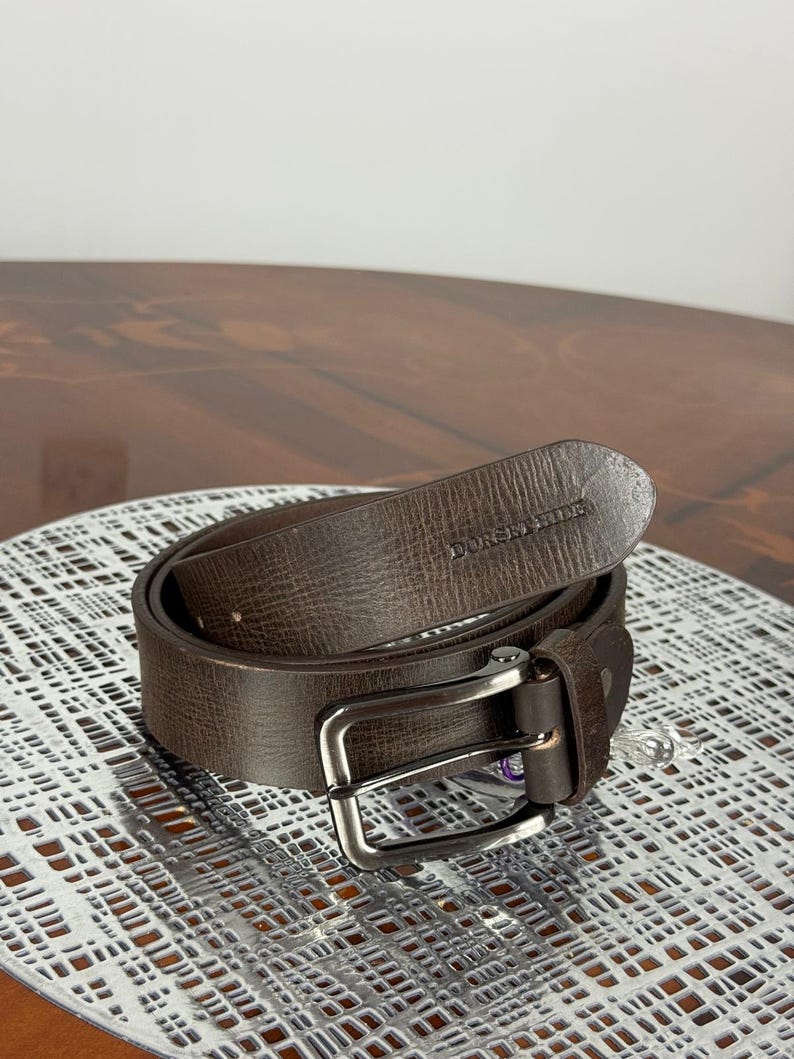 May include: A brown leather belt with a silver-colored buckle. The belt is rolled up and has the brand name "DUNSHILL" embossed on the leather. The belt is on a white patterned surface.