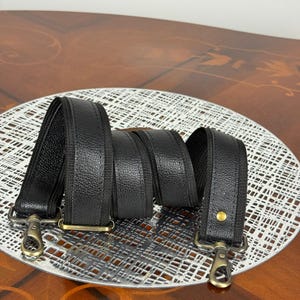 Puede incluir: Correa de bolso de cuero negro con herrajes metálicos dorados. La correa está hecha de un material de cuero texturizado y presenta un diseño resistente. La correa tiene un clip en cada extremo para una fácil fijación a un bolso.