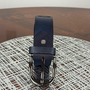 May include: A navy blue leather belt with a silver-colored buckle. The belt is shown on a textured surface. The buckle is a classic design with a single prong. The belt is a standard width.