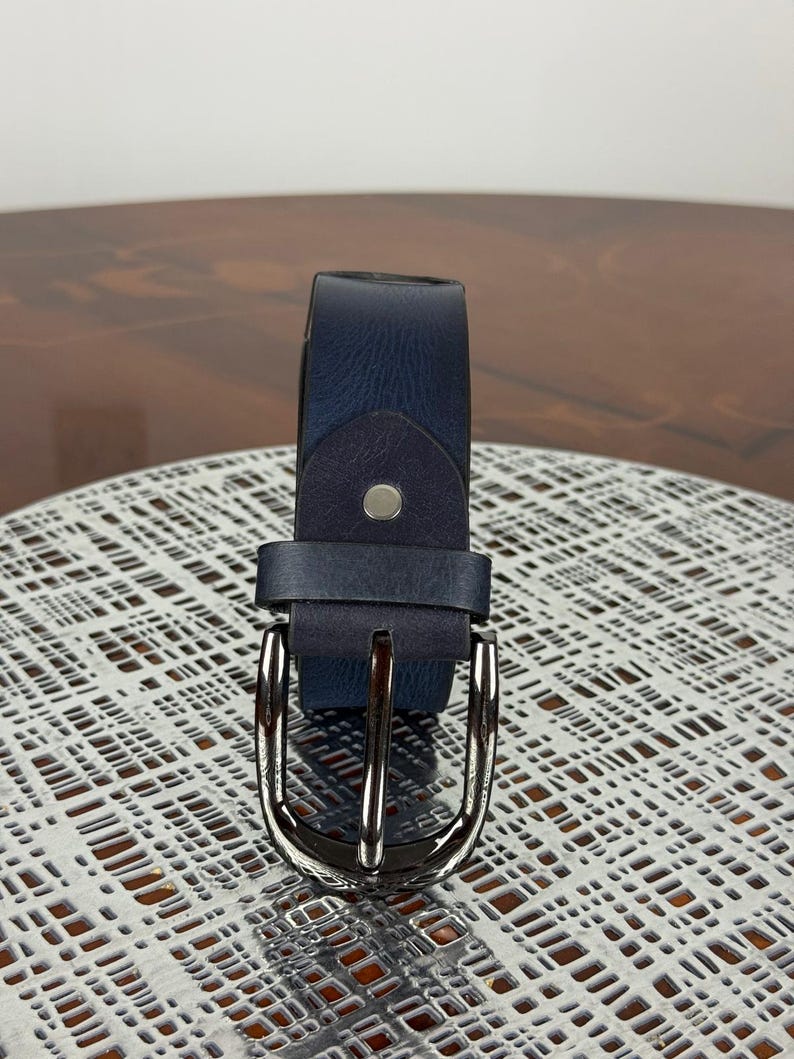 May include: A navy blue leather belt with a silver-colored buckle. The belt is shown on a textured surface. The buckle is a classic design with a single prong. The belt is a standard width.