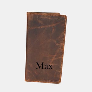 Puede incluir: Cartera de cuero marrón con el nombre "Max" grabado en la parte delantera. La cartera tiene un acabado envejecido y costuras visibles en los bordes. La marca "DORSET HIDE" está impresa en la parte inferior.