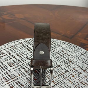 May include: A brown leather belt with a silver-colored buckle. The belt is made of a dark brown leather material and has a simple, classic design. The buckle is rectangular and has a shiny finish. The belt is displayed on a white surface.