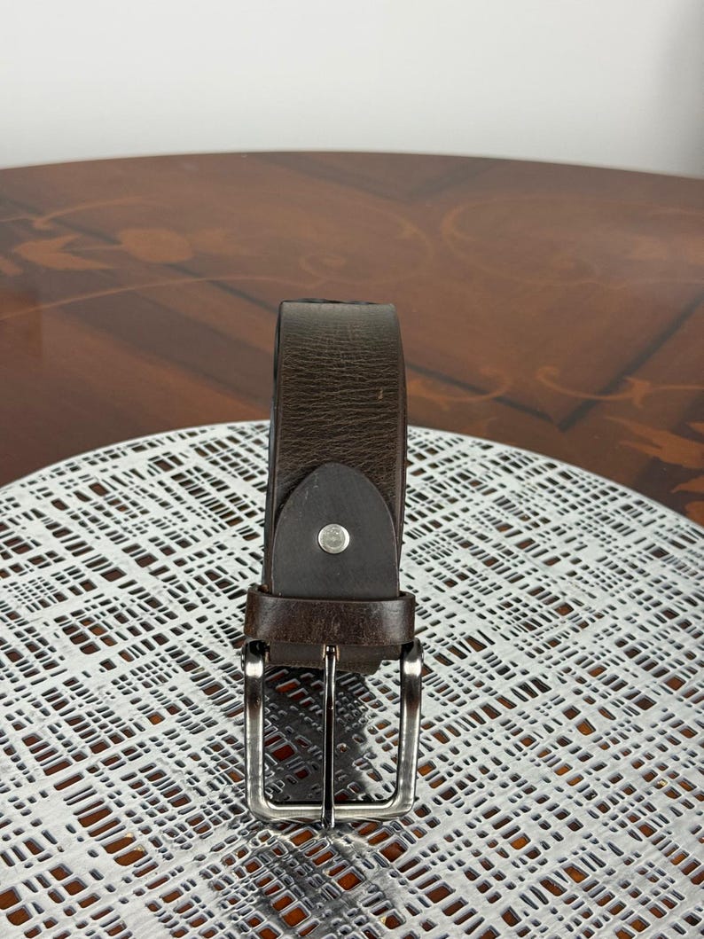 May include: A brown leather belt with a silver-colored buckle. The belt is made of a dark brown leather material and has a simple, classic design. The buckle is rectangular and has a shiny finish. The belt is displayed on a white surface.