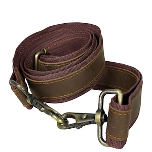 May include: A brown and burgundy bag strap with metal fixings. The strap is made of a woven burgundy material with brown leather accents and stitching. The metal fixings have an antique brass finish.