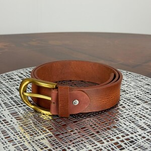 May include: A rolled brown leather belt with a gold-colored buckle. The belt has a classic design and a textured surface. The buckle is rectangular with rounded edges. The belt is a classic accessory.
