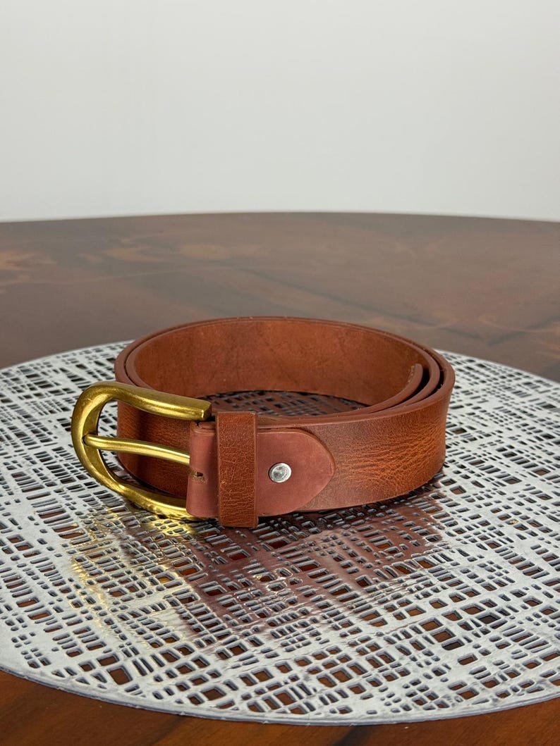 May include: A rolled brown leather belt with a gold-colored buckle. The belt has a classic design and a textured surface. The buckle is rectangular with rounded edges. The belt is a classic accessory.