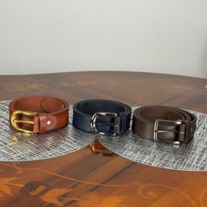 May include: Three leather belts in shades of brown and navy blue are displayed. The belts have silver and gold-colored buckles. The belts are arranged on a decorative surface.