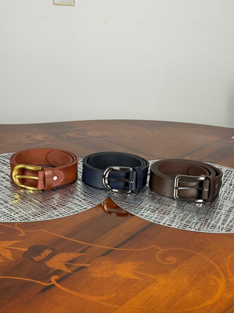 May include: Three leather belts in shades of brown and navy blue are displayed. The belts have silver and gold-colored buckles. The belts are arranged on a decorative surface.