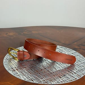 May include: A brown leather belt with a gold-colored buckle. The belt is displayed on a decorative silver-colored plate. The belt has a rectangular end with the word "FOREVER" embossed on it.