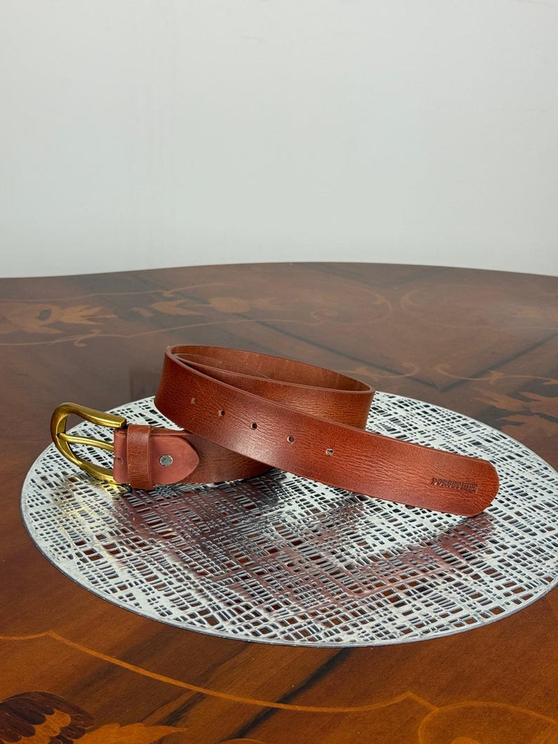 May include: A brown leather belt with a gold-colored buckle. The belt is displayed on a decorative silver-colored plate. The belt has a rectangular end with the word "FOREVER" embossed on it.