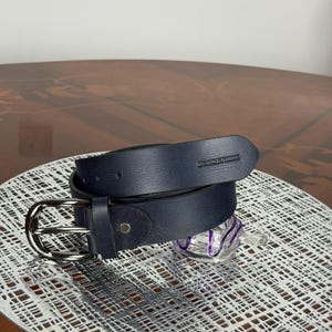 May include: A navy blue leather belt with a silver-colored buckle. The belt is rolled up and has the brand name "DORSERHILL" embossed on the end. The belt is on a white patterned surface.
