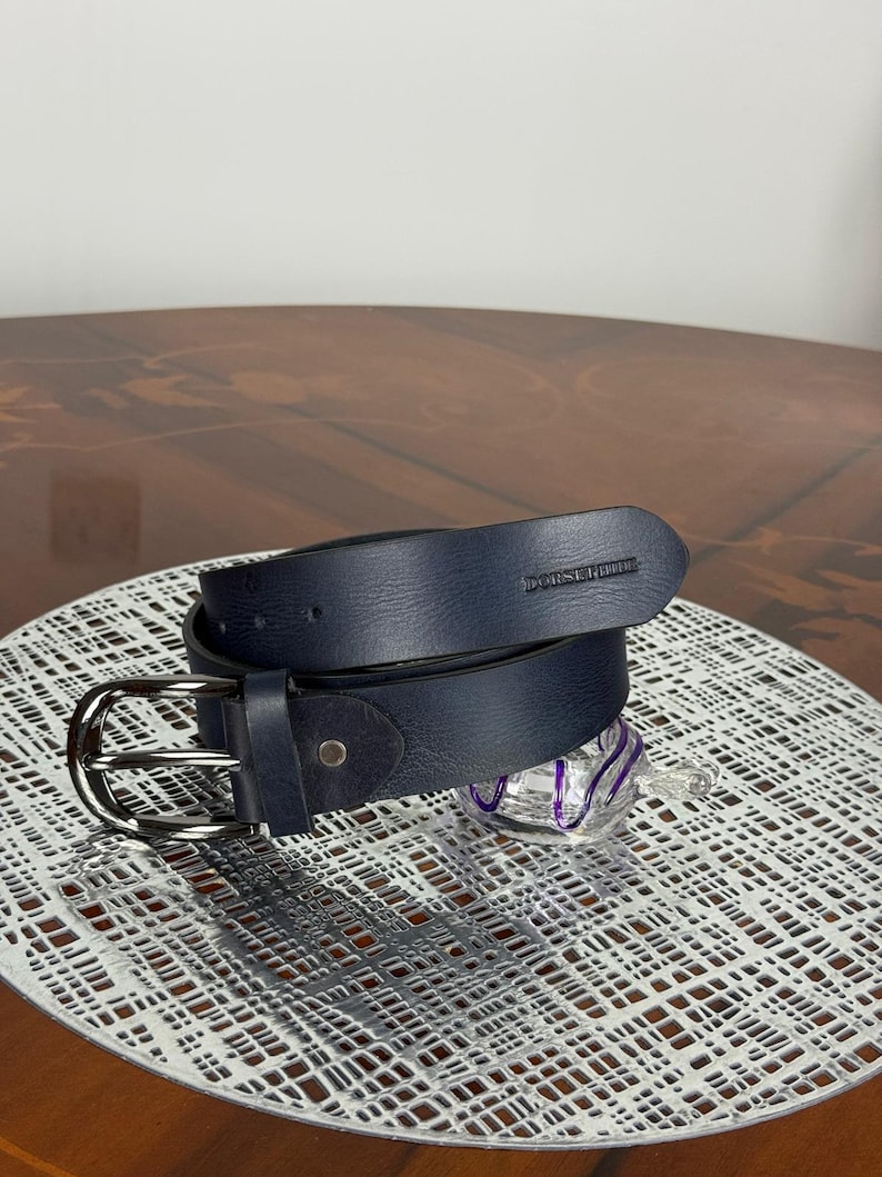 May include: A navy blue leather belt with a silver-colored buckle. The belt is rolled up and has the brand name "DORSERHILL" embossed on the end. The belt is on a white patterned surface.