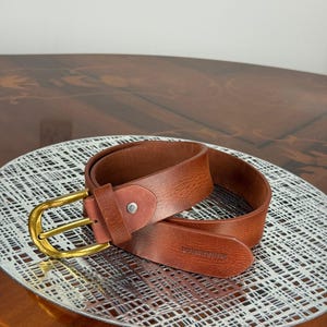 May include: A brown leather belt with a gold-colored buckle. The belt is rolled up on a silver-colored, patterned surface. The belt has the text "DORSEWOOD" embossed on the leather.