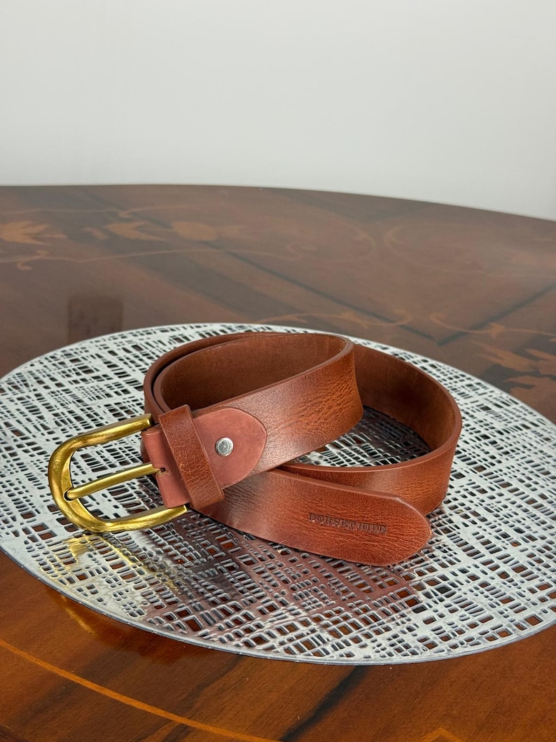 May include: A brown leather belt with a gold-colored buckle. The belt is rolled up on a silver-colored, patterned surface. The belt has the text "DORSEWOOD" embossed on the leather.