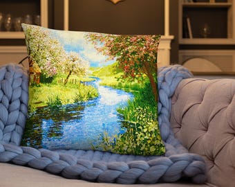 May include: A decorative pillow featuring a scenic landscape with a river flowing through a lush green valley. The pillow showcases a vibrant painting with trees in full bloom, reflecting in the blue water. The pillow is placed on a light gray knitted blanket.