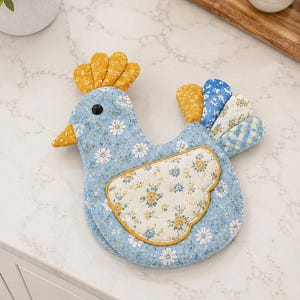 May include: A fabric chicken-shaped pot holder featuring a light blue base with a white daisy floral pattern. It has a yellow patterned comb and beak, a black eye, and a cream-colored wing with blue and yellow flowers. The tail feathers are multicolored.
