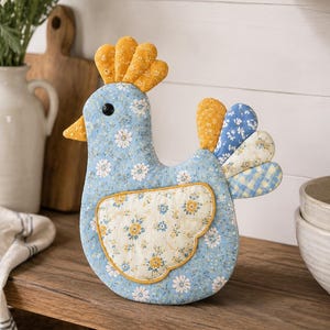 May include: A stuffed fabric chicken ornament featuring a light blue floral print body, a yellow comb, and a cream-colored wing with yellow and blue floral patterns. The tail consists of layered fabric segments in yellow, blue, and cream floral and plaid prints.