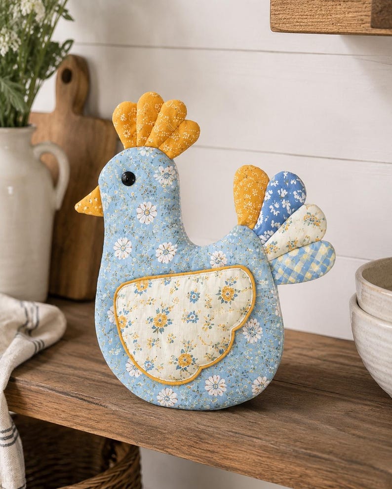 May include: A stuffed fabric chicken ornament featuring a light blue floral print body, a yellow comb, and a cream-colored wing with yellow and blue floral patterns. The tail consists of layered fabric segments in yellow, blue, and cream floral and plaid prints.