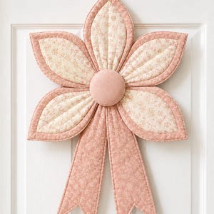 May include: A quilted fabric flower wall hanging in shades of dusty rose and cream. The flower features five petals with a cream floral print center and solid dusty rose borders, a central dusty rose button, and two long dusty rose floral ribbons.