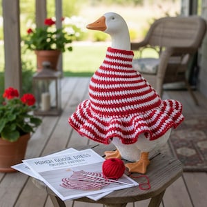 May include: A white goose statue wearing a red and white striped crochet dress. The goose is standing on a wooden table with a ball of red yarn, a crochet hook, and a pattern titled "Porch Goose Dress Crochet Pattern."