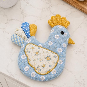 May include: A fabric chicken-shaped potholder featuring a light blue base with a white floral pattern. It has a yellow comb, a yellow beak, and a black eye. The wing is a cream-colored fabric with yellow and blue flowers, and the tail feathers are blue, yellow, and white with various patterns.