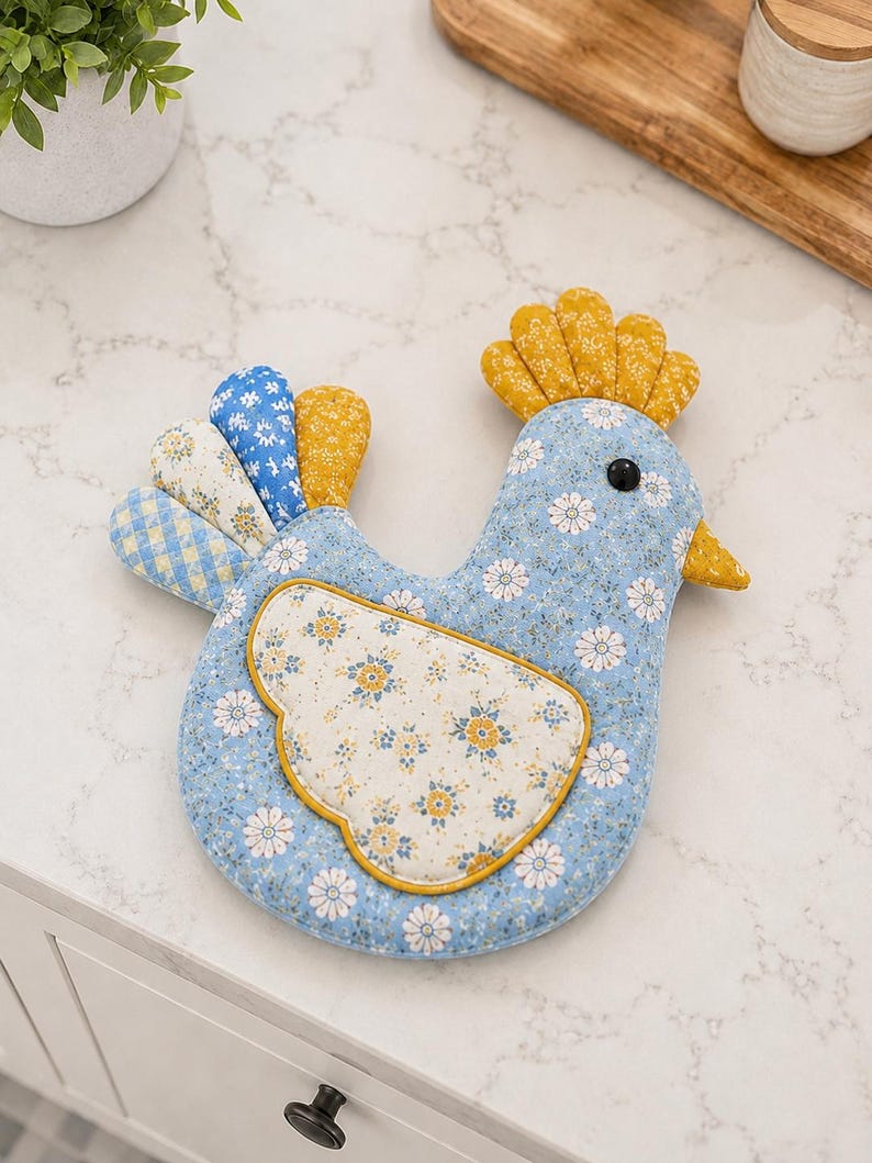 May include: A fabric chicken-shaped potholder featuring a light blue base with a white floral pattern. It has a yellow comb, a yellow beak, and a black eye. The wing is a cream-colored fabric with yellow and blue flowers, and the tail feathers are blue, yellow, and white with various patterns.