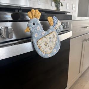 May include: A chicken-shaped fabric oven mitt featuring a light blue base with a white and yellow floral pattern. The comb and tail feathers are made of yellow and blue patterned fabric, and the wing is a cream-colored patch with floral details.