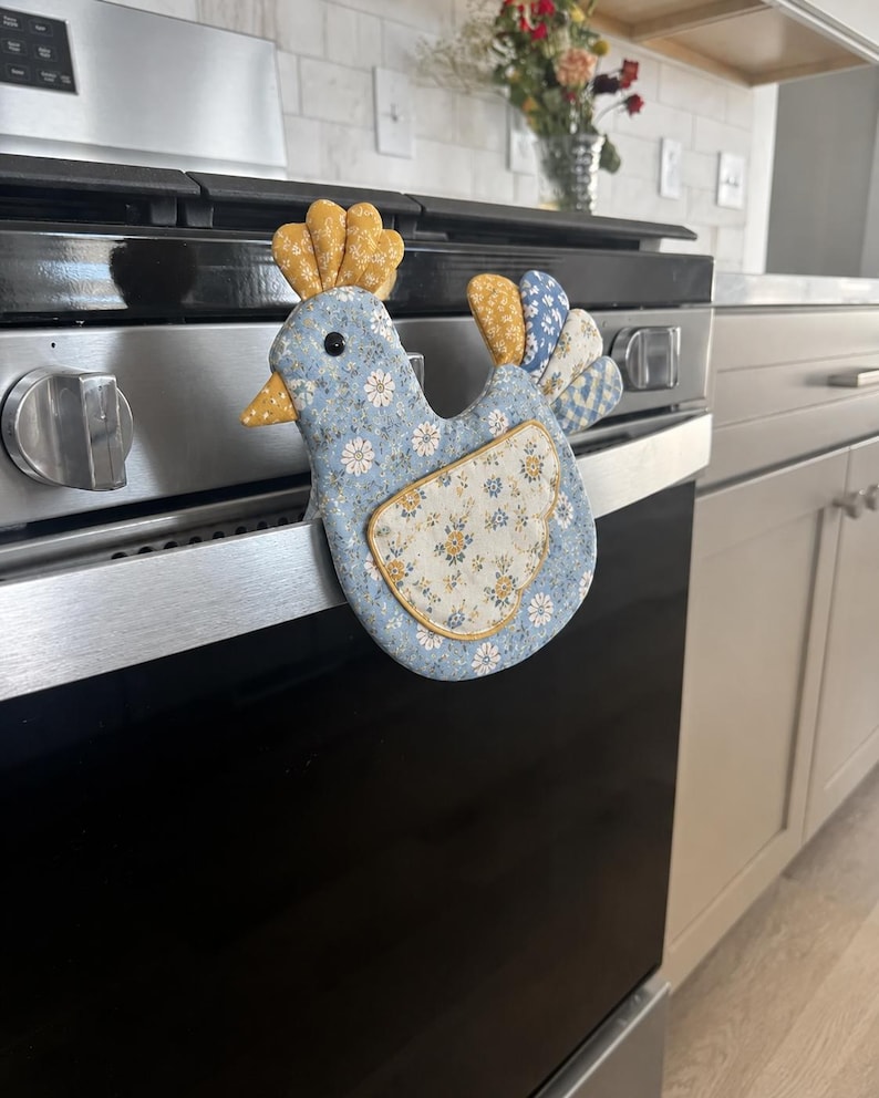 May include: A chicken-shaped fabric oven mitt featuring a light blue base with a white and yellow floral pattern. The comb and tail feathers are made of yellow and blue patterned fabric, and the wing is a cream-colored patch with floral details.