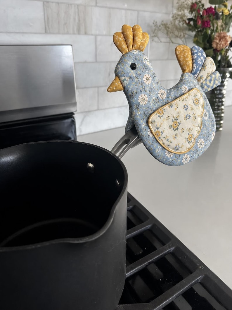 May include: A fabric pot handle cover shaped like a chicken with a blue floral pattern, a yellow comb, and a yellow beak. The cover is attached to the handle of a black saucepan, which is resting on a black stovetop burner in a kitchen setting.