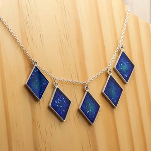 May include: A silver chain necklace featuring six diamond-shaped pendants. Each pendant displays a dark blue design with white speckles, reminiscent of a starry night. The necklace is presented on a wooden jewellery stand.