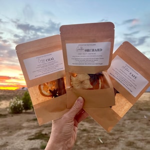May include: Three brown paper pouches of stovetop potpourri labeled Cozy Chai, Golden Orchard, and Autumn Cafe. Each pouch features a clear window revealing dried orange slices, star anise, and other botanicals inside the packaging.