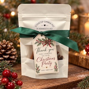 May include: A white pouch labeled Stovetop Simmer Pot by Ironwood & Lace, tied with a dark green ribbon. A tag attached to the ribbon reads Thank you for coming to our Christmas Party, The Smiths, decorated with pine branches and red berries.