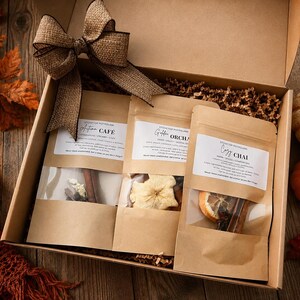 May include: A cardboard gift box containing three kraft paper pouches of stovetop potpourri labeled Autumn Cafe, Golden Orchard, and Cozy Chai. The pouches feature clear windows revealing dried fruit and spices, topped with a large burlap bow.
