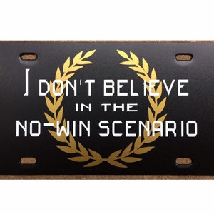 May include: Black license plate with gold laurel wreath and white text that reads "I don't believe in the no-win scenario."
