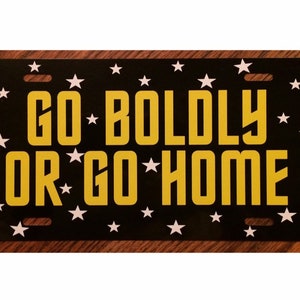 May include: Black metal license plate with white stars and yellow text that reads "GO BOLDLY OR GO HOME".