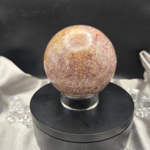 Può includere: Una gemma sferica lucidata con un aspetto screziato, che mostra una miscela di tonalità marrone-rossastro e dorato. La sfera è esposta su un piedistallo nero, creando un contrasto sorprendente. La superficie della gemma ha un aspetto strutturato.